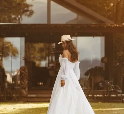 Una novia con un vestido blanco y un sombrero se encuentra al aire libre junto a un edificio rústico.