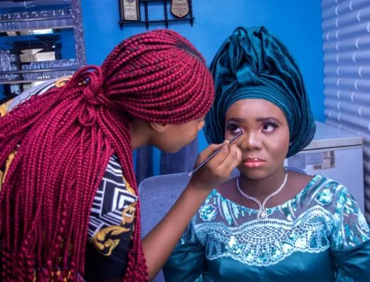 Una mujer nigeriana vestida con traje tradicional recibiendo maquillaje de un artista de maquillaje en interiores.