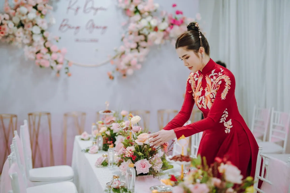 Una mujer asiática con un vestido tradicional rojo organiza un centro de mesa floral en un elegante evento interior (imagen de referencia)