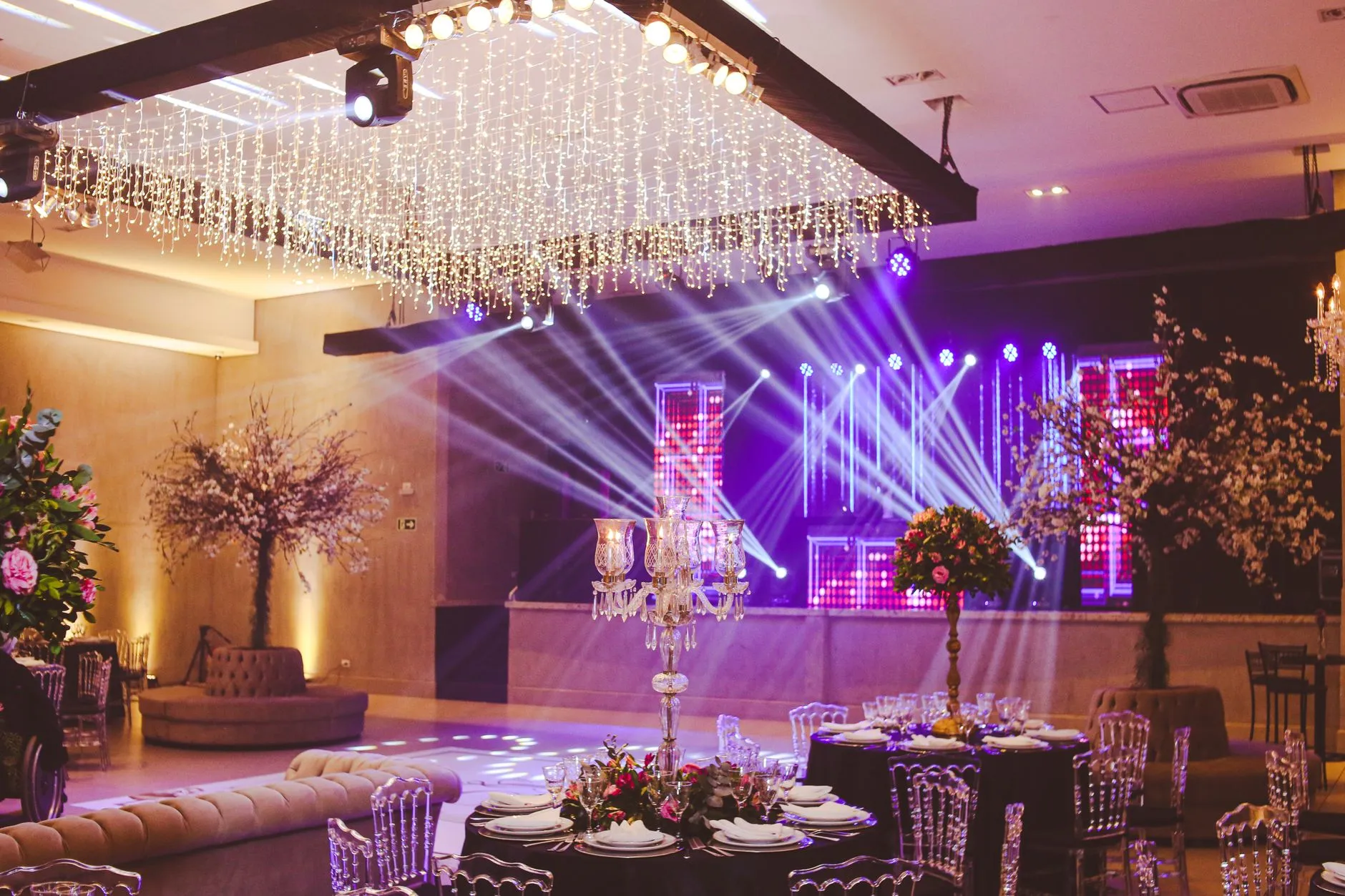 Impresionante montaje de salón decorado con candelabros de cristal, arreglos florales y vibrante iluminación en el escenario.