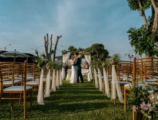 Una romántica decoración de boda al aire libre con una pareja abrazándose entre sillas decoradas y vegetación.