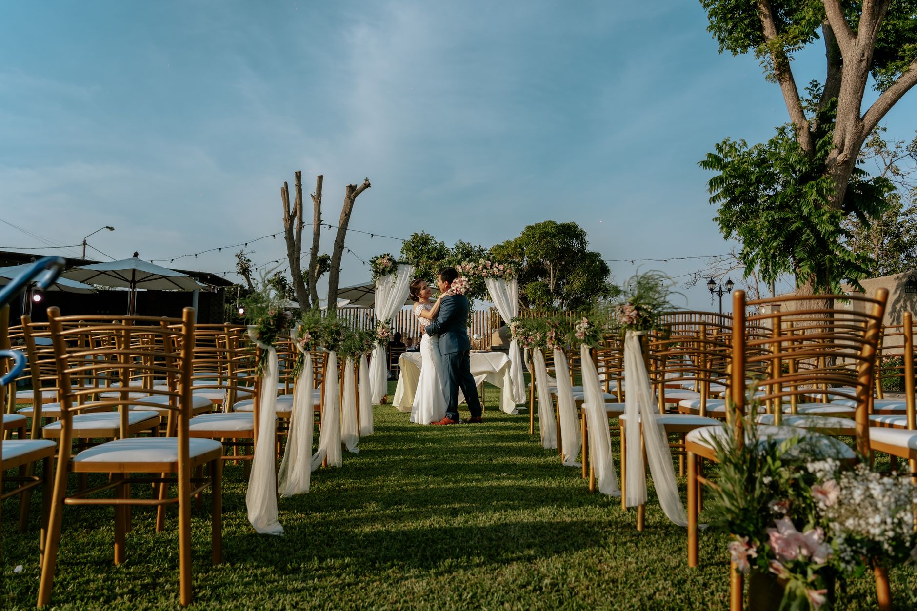 Una romántica decoración de boda al aire libre con una pareja abrazándose entre sillas decoradas y vegetación.