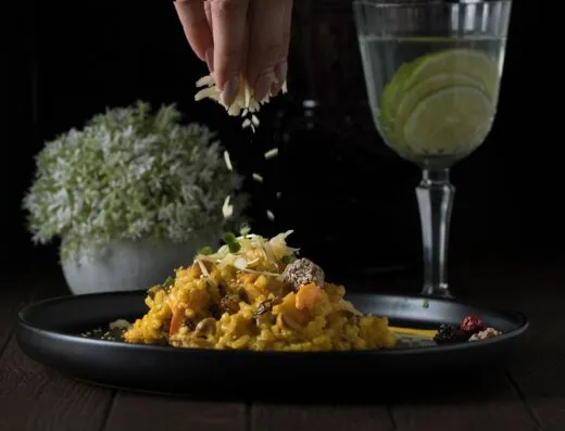 Delicioso risotto siendo decorado con hierbas junto a una refrescante bebida de lima sobre un fondo oscuro.