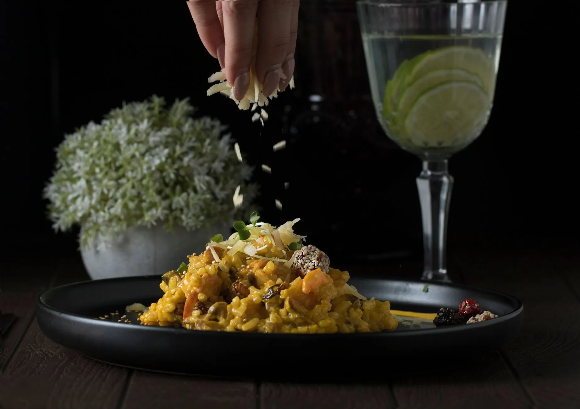 Delicioso risotto siendo decorado con hierbas junto a una refrescante bebida de lima sobre un fondo oscuro.