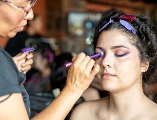 Una mujer recibe un intrincado maquillaje de Catrina mexicana en el interior, centrándose en el arte colorido de los ojos.