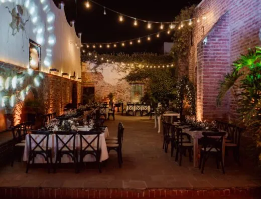 Encantador entorno para eventos al aire libre por la noche, con mesas con una decoración elegante bajo cálidas luces de cadena.