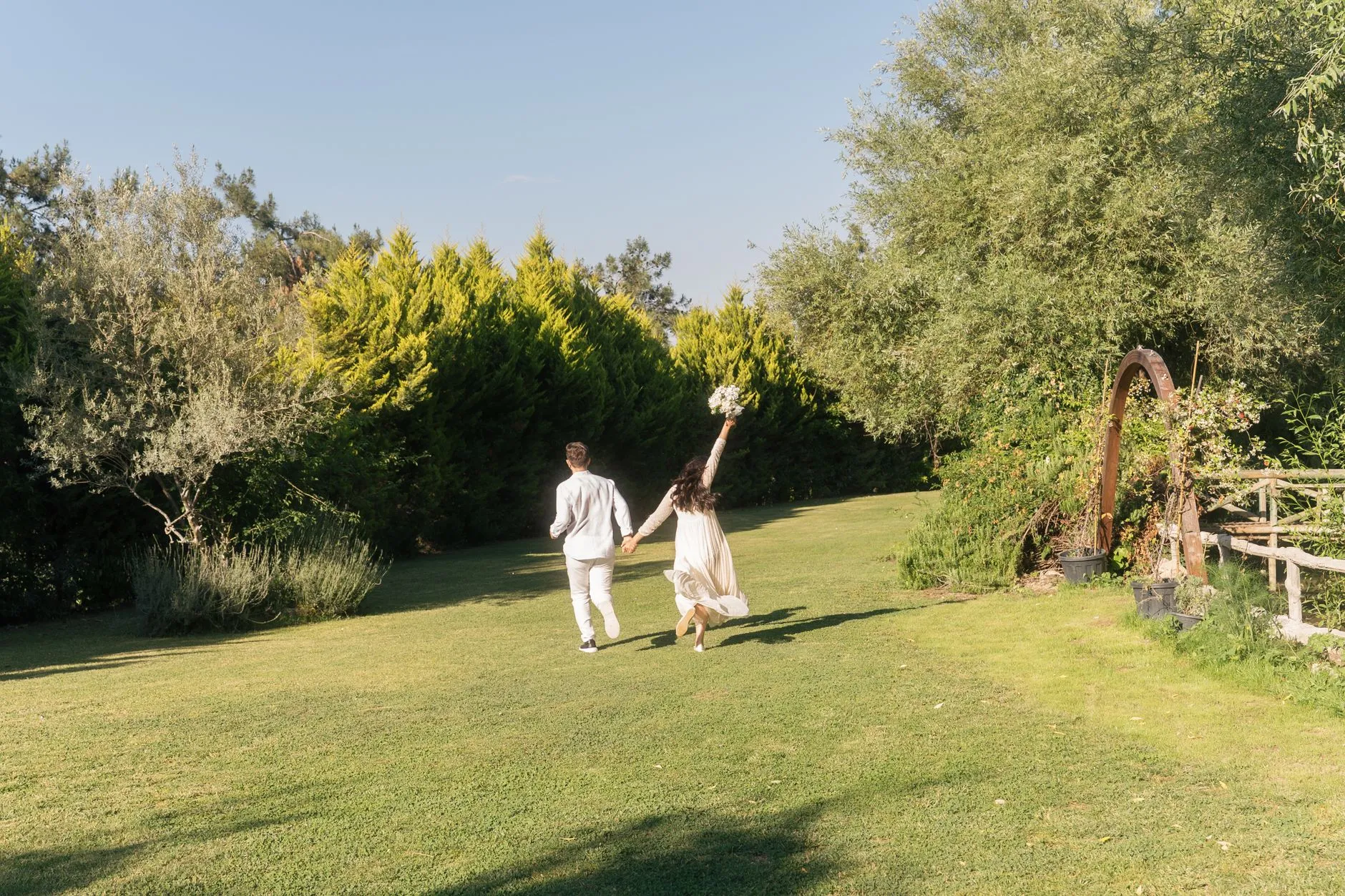 Una pareja vestida de novia corriendo alegremente por un jardín soleado.