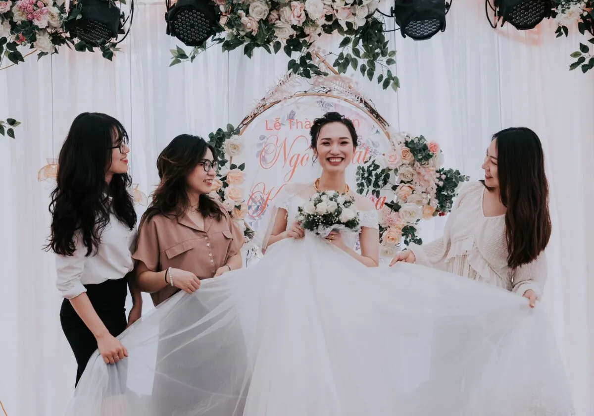 Novia alegre con damas de honor sosteniendo su vestido, celebrando una ceremonia de boda en interiores (imagen de referencia)