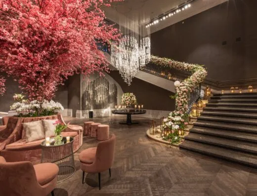 Impresionante vestíbulo de hotel adornado con arreglos florales rosas y una elegante zona de estar bajo una gran escalera.