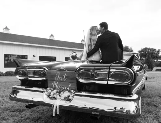 Los recién casados comparten un beso en un convertible clásico con un cartel de 'Recién Casados'.