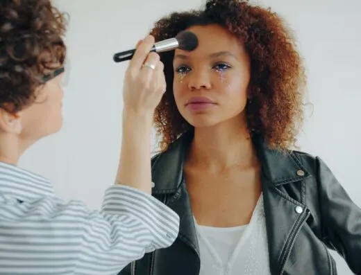 Maquilladora profesional aplicando maquillaje a una joven en el interior de un edificio.