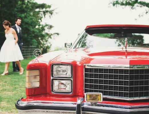 Un coche clásico rojo estacionado al aire libre en una boda con una novia y un novio al fondo.