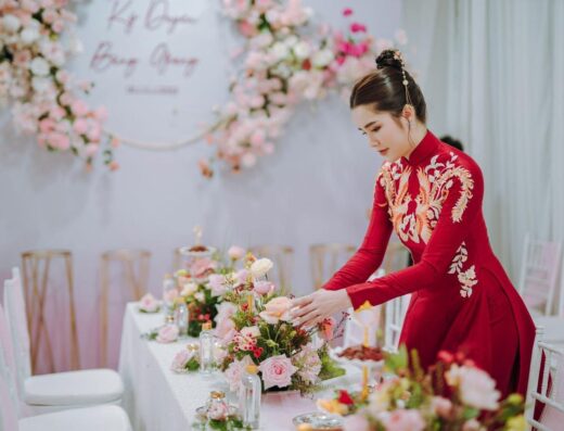 Una mujer asiática con un vestido tradicional rojo organiza un centro de mesa floral en un elegante evento interior.