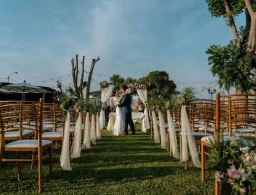 Una romántica decoración de boda al aire libre con una pareja abrazándose entre sillas decoradas y vegetación.