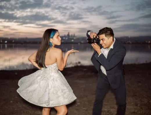 Una mujer con un vestido posa para un hombre con una cámara junto al lago al atardecer.
