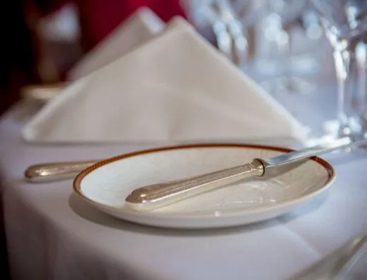 Primer plano de una configuración de mesa formal con cubiertos, un plato y una servilleta doblada sobre un mantel blanco.