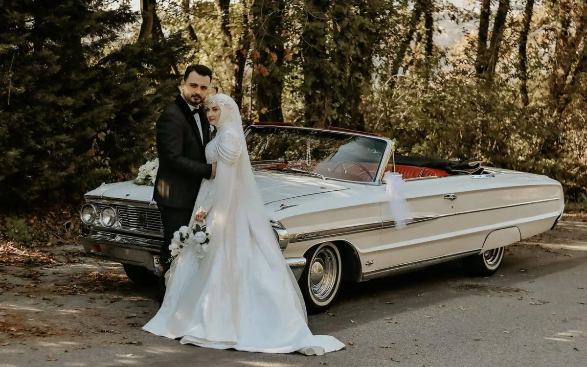 La novia y el novio posan románticamente junto a un convertible clásico en un hermoso entorno al aire libre (imagen de referencia)