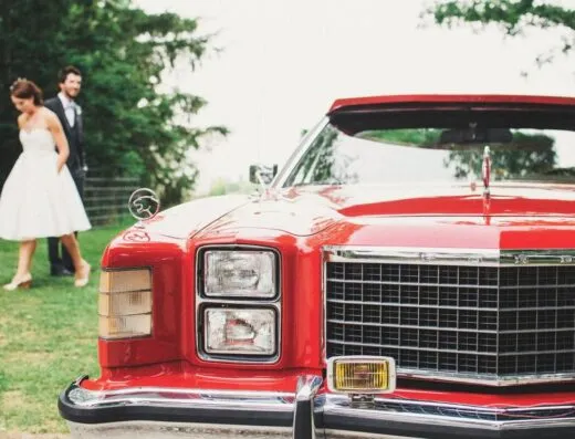 Un coche clásico rojo estacionado al aire libre en una boda con una novia y un novio al fondo.