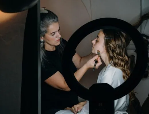 Una maquilladora aplica cosméticos hábilmente a una mujer bajo una luz anular en un entorno interior.