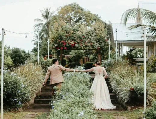 Una pareja de recién casados de la mano en un jardín frondoso, capturada desde atrás.