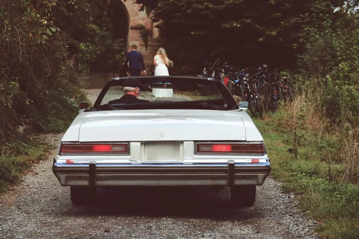 Un coche vintage estacionado en un camino de grava, con una novia y un novio caminando adelante (imagen de referencia)