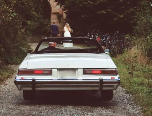 Un coche vintage estacionado en un camino de grava, con una novia y un novio caminando adelante.
