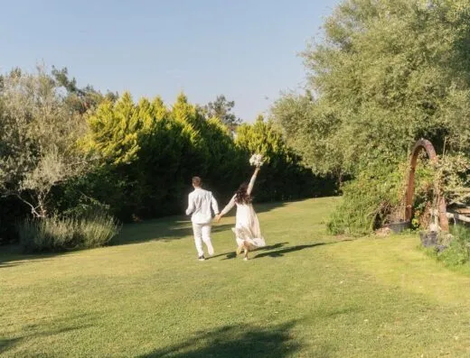 Una pareja vestida de novia corriendo alegremente por un jardín soleado.