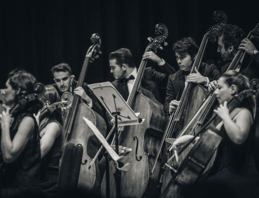 Foto en blanco y negro de una orquesta clásica actuando en vivo, centrada en músicos de cuerdas.