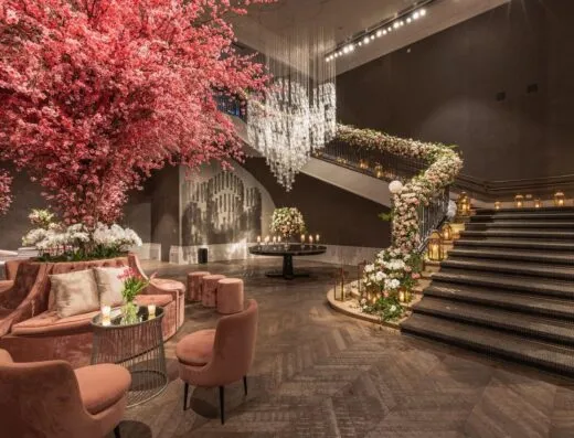 Impresionante vestíbulo de hotel adornado con arreglos florales rosas y una elegante zona de estar bajo una gran escalera.