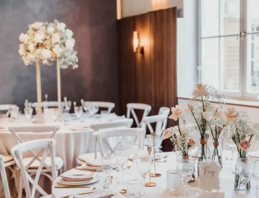 Montaje de boda lujoso con manteles blancos, centros de flores y vajilla elegante en un lugar brillante.