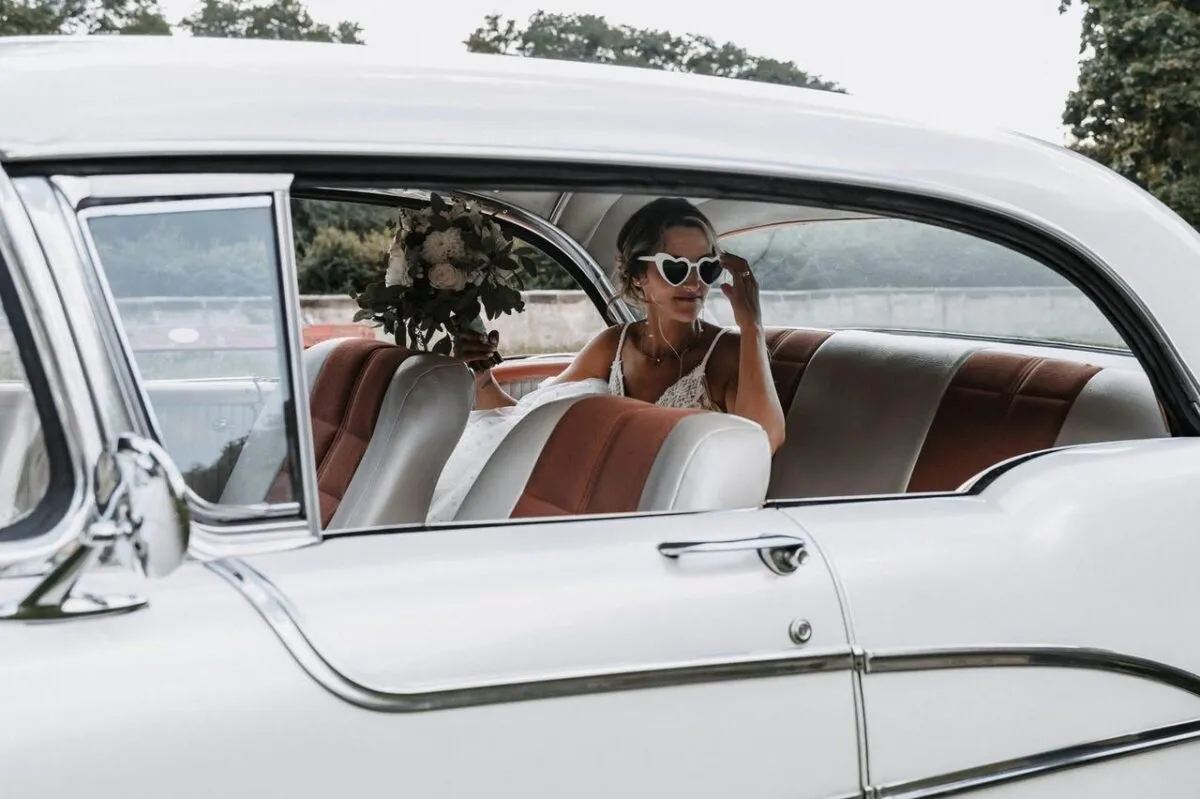 Novia sentada en un coche vintage, usando gafas en forma de corazón y sosteniendo un ramo. Perfecto para temas de boda (imagen de referencia)