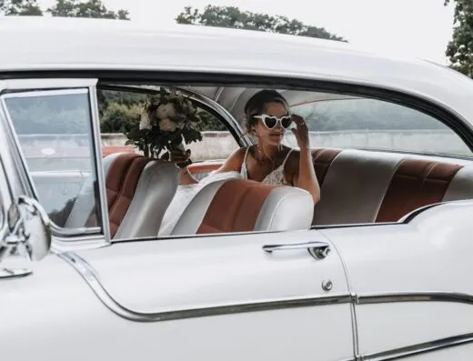 Novia sentada en un coche vintage, usando gafas en forma de corazón y sosteniendo un ramo. Perfecto para temas de boda.
