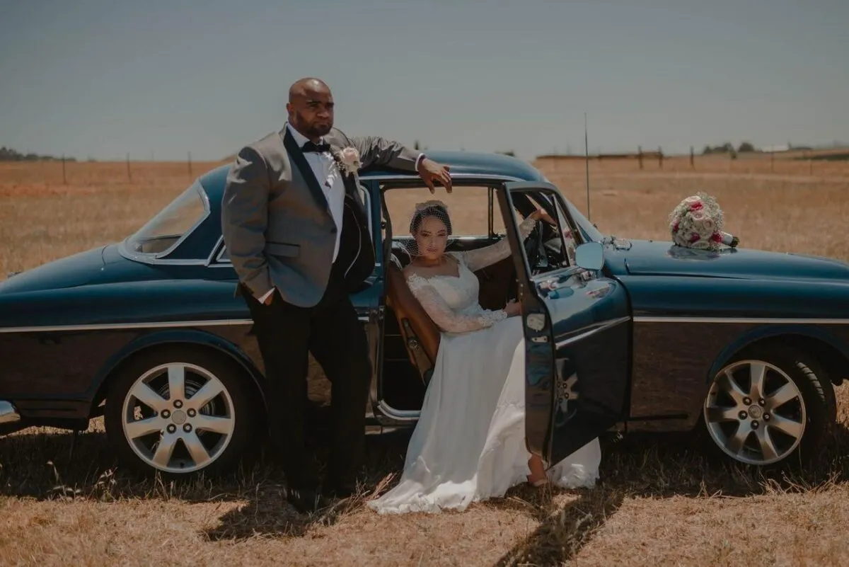 Pareja elegante en traje de boda posando con un coche clásico en un campo abierto (imagen de referencia)