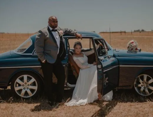 Pareja elegante en traje de boda posando con un coche clásico en un campo abierto.