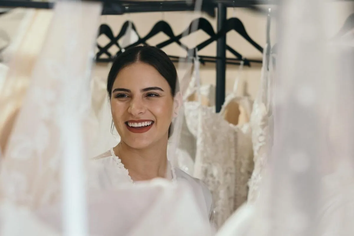 Una novia alegre sonríe mientras está rodeada de hermosos vestidos de novia en perchas (imagen de referencia)