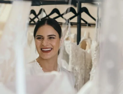 Una novia alegre sonríe mientras está rodeada de hermosos vestidos de novia en perchas.