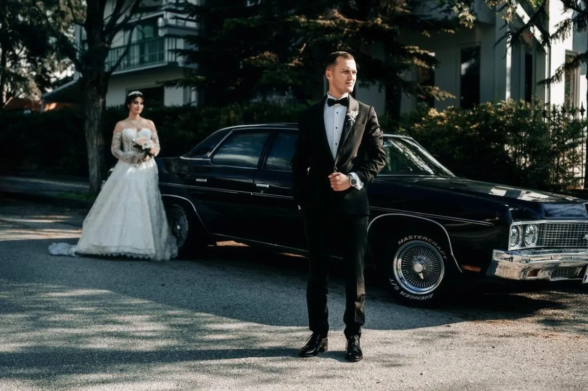 Una novia y un novio vestidos de gala posan junto a un coche clásico en un día soleado (imagen de referencia)