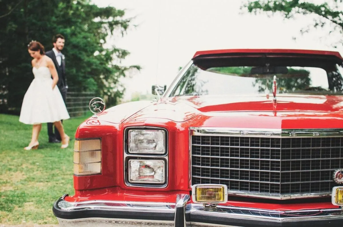Un coche clásico rojo estacionado al aire libre en una boda con una novia y un novio al fondo (imagen de referencia)