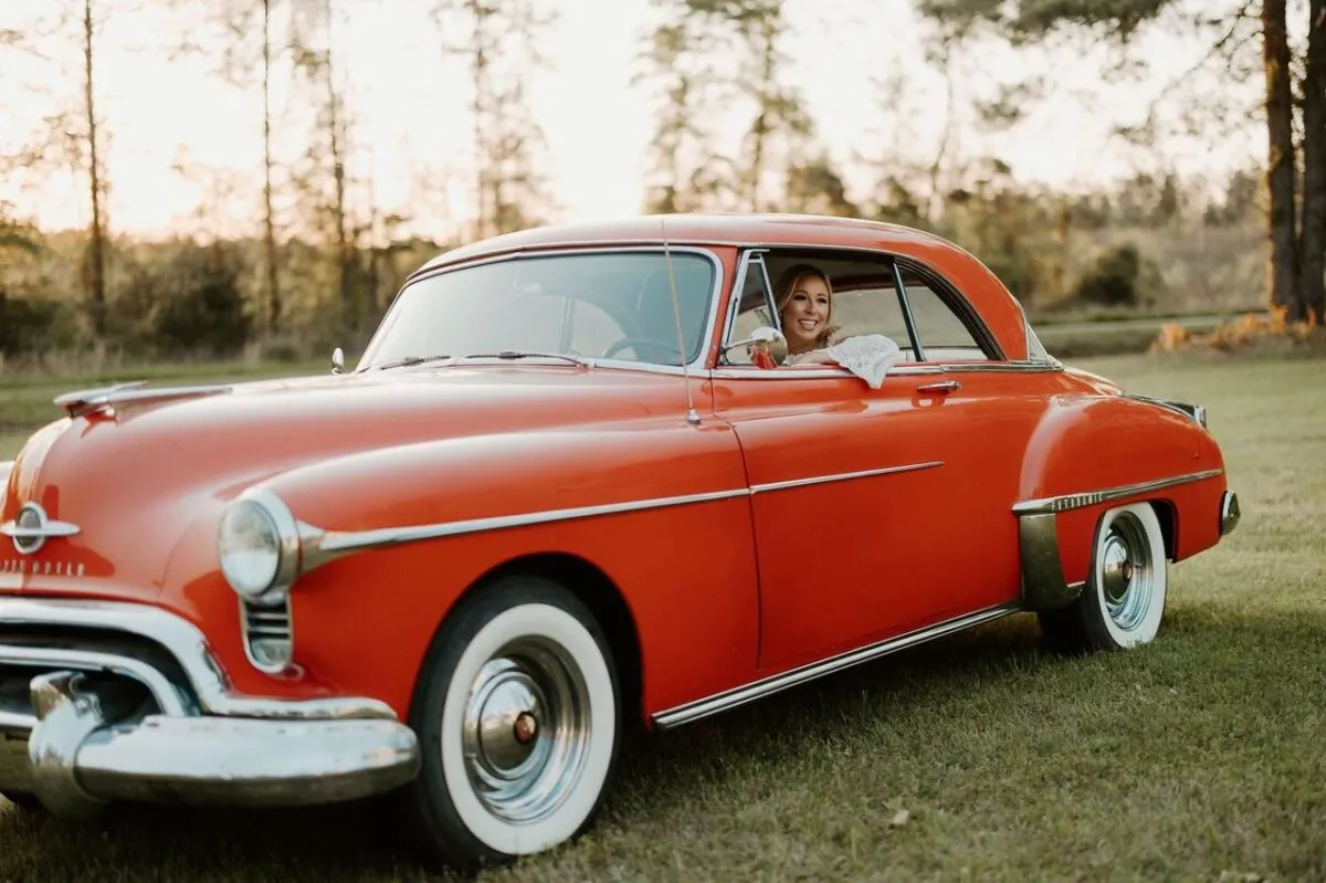 Una mujer sonriente conduce un clásico coche vintage rojo en un día soleado al aire libre (imagen de referencia)
