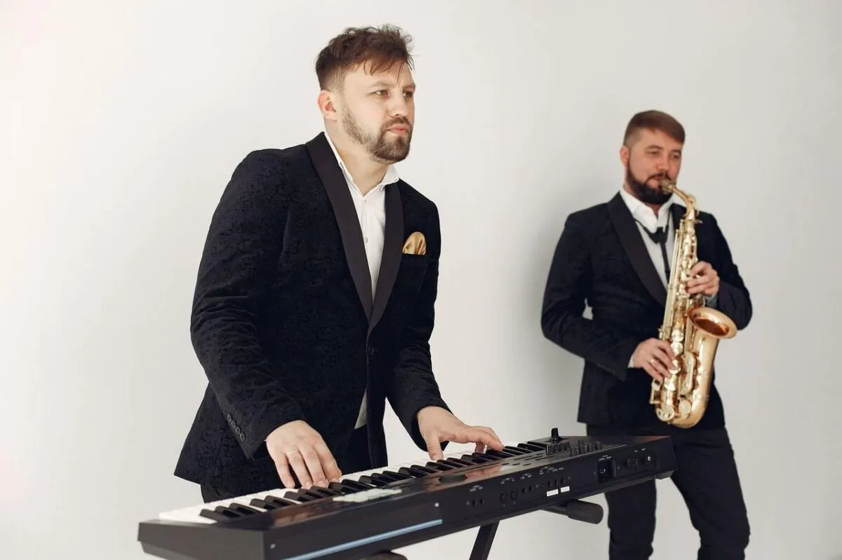 Dos músicos hombres en trajes tocando un teclado y un saxofón en un estudio (imagen de referencia)
