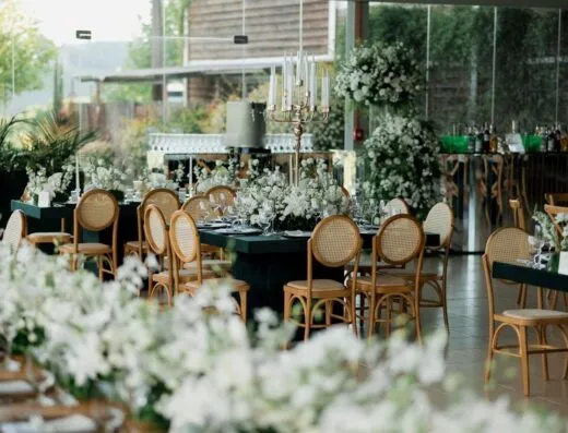 Decoración de evento chic con arreglos florales y sillas de madera en una brillante sala soleada.